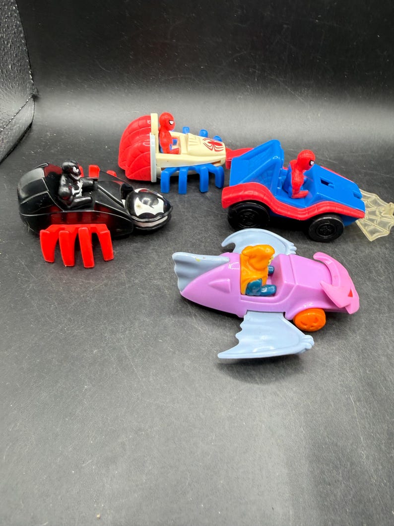 May include: Four toy vehicles featuring Spider-Man characters. The toys include a black car with red accents, a red and blue car, a red and blue vehicle with a trailer, and a pink and blue aircraft. Each vehicle has a Spider-Man figure.