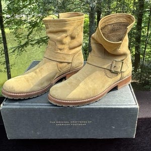 Könnte beinhalten: Hellbraune Wildleder-Ankle Boots mit Schnallenverschluss und kleinem Absatz. Sie stehen auf einer grauen Box mit der Aufschrift "THE ORIGINAL CRAFTSMEN OF AMERICAN FOOTWEAR". Die Stiefel haben einen Used-Look.