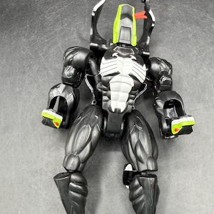 May include: Black and white action figure with green and red details. The figure has a muscular physique, a white chest emblem, and claw-like feet. Two long, curved appendages extend from the head.