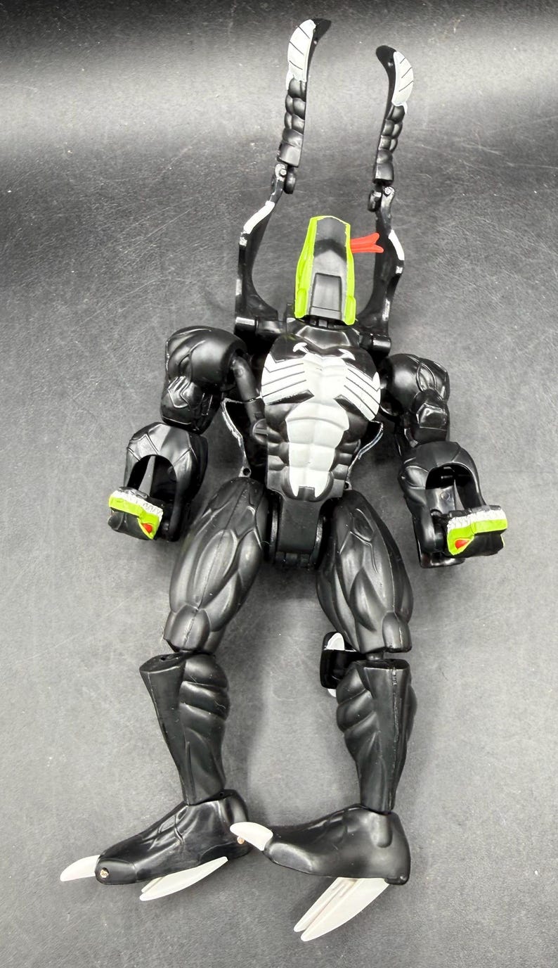 May include: Black and white action figure with green and red details. The figure has a muscular physique, a white chest emblem, and claw-like feet. Two long, curved appendages extend from the head.