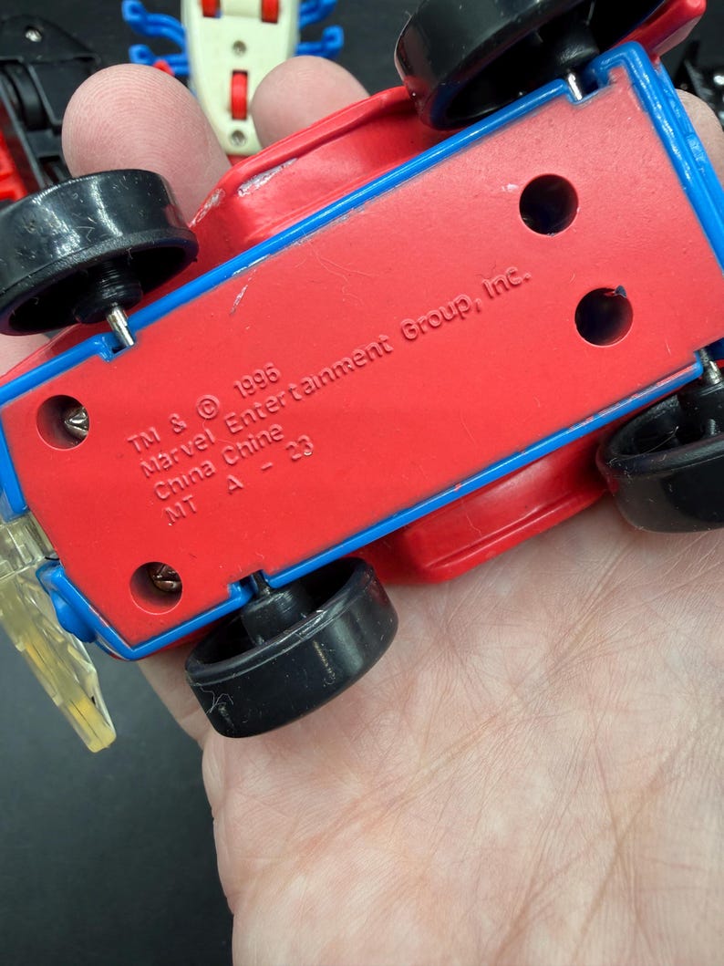 May include: A red and blue toy car with black wheels. The underside of the car features the text "TM & &copy; 1996 Marvel Entertainment Group, Inc. China Chine MT A - 23".