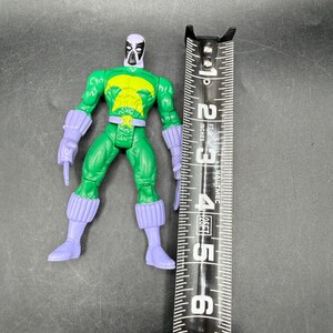 May include: A green action figure with purple gloves, boots, and a mask. The figure has a yellow chest emblem and is approximately 13 cm tall. A black and silver measuring tape is next to the figure.