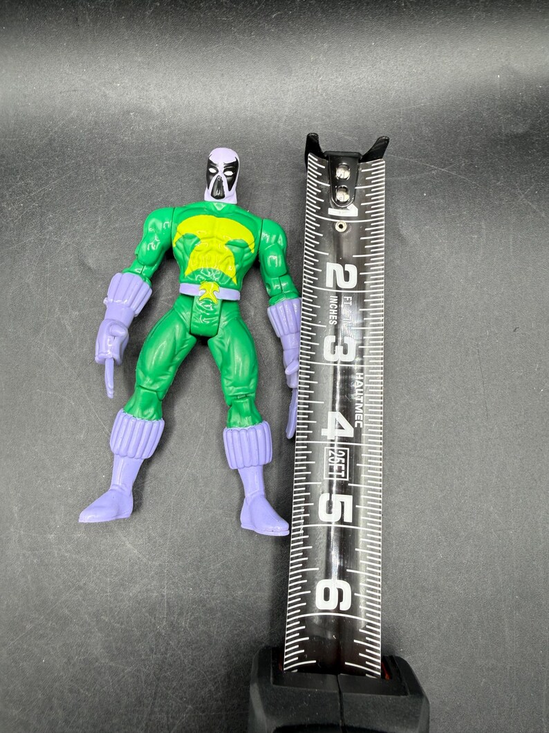 May include: A green action figure with purple gloves, boots, and a mask. The figure has a yellow chest emblem and is approximately 13 cm tall. A black and silver measuring tape is next to the figure.