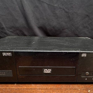 May include: A black DVD player with a silver faceplate and the text "DVD" and "Dolby Digital" on it. The player is sitting on a wooden surface.