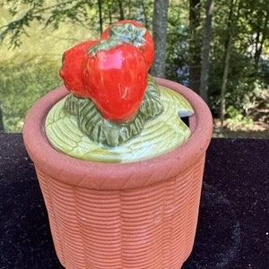 Peut inclure: Un pot à sucre en céramique avec un couvercle en forme de panier de fraises. Le couvercle est décoré de deux fraises rouges avec des feuilles vertes.