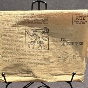 May include: A vintage envelope with a brown background and white text. The envelope is addressed to a subscriber and includes a coupon for a subscription to "The Workbasket". The envelope also features a stamp that says "U.S. Postage Paid".
