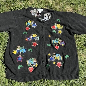 以下が含まれることがあります： ボタン留めの黒いカーディガン。カラフルな刺繍の天使と星が施されています。