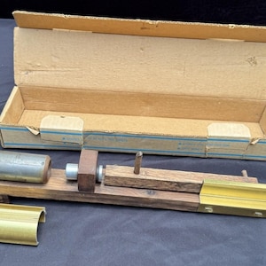 Könnte beinhalten: Ein Holzwerkzeug mit einem Metallzylinder, einem quadratischen Block und einem goldfarbenen Metallaufsatz, begleitet von einem separaten goldfarbenen, gebogenen Metallteil und einem offenen rechteckigen Karton mit blauen Linien.