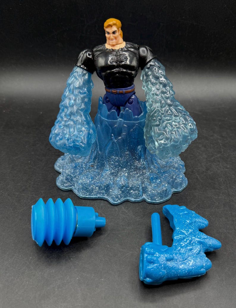 May include: A superhero action figure with a black top, blue pants, and water-like arms and base. Includes two blue accessories. The figure has blonde hair and a determined expression.