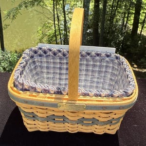 Peut inclure: Un panier tressé rectangulaire avec une anse en bois verticale, composé de brins naturels et bleu-gris. Il comprend une doublure en tissu à carreaux bleus et blancs et un insert en plastique transparent. Une plaque en laiton indique Longaberger 1996.