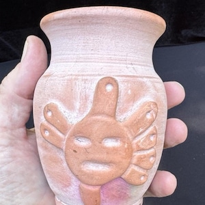 May include: A small, hand-crafted terracotta vase with a sun-like face design. The vase is a light pink color with a darker terracotta-colored sun design. The vase is held in a hand.