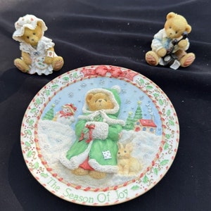 Könnte beinhalten: Ein weißer Keramikteller mit einer Winterszene, die einen Bären zeigt, der einen grünen Mantel trägt und ein Geschenk hält. Der Teller hat einen roten und weißen Zuckerstangenrand und den Text "The Season of Joy".