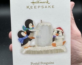 May include: Hallmark Keepsake Christmas tree ornament featuring three penguin figures. One penguin wears a Santa hat, another holds a letter, and the third holds a sign. The ornament is packaged in a white box with the words "Postal Penguins".
