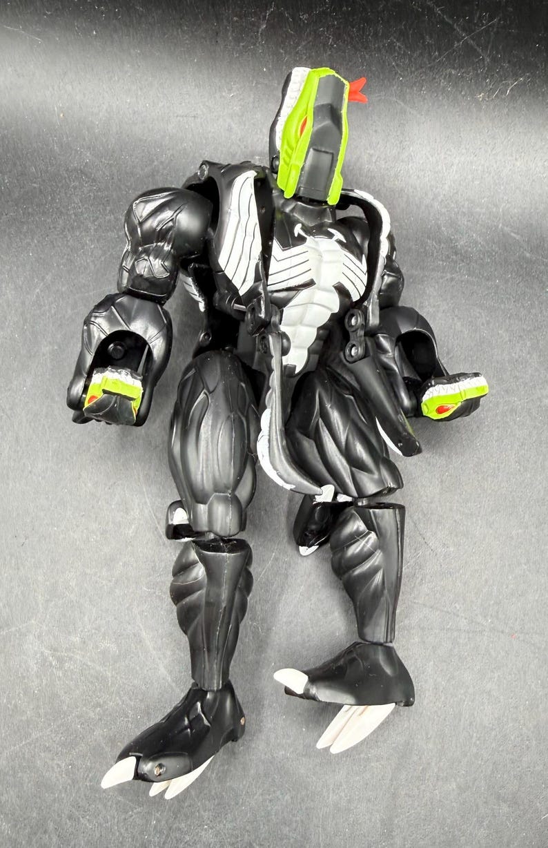 May include: A black and white action figure with a green and red head. The figure has a muscular build with detailed textures and accents. The figure's hands and feet have green and white details.