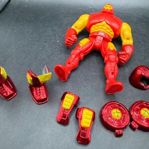 May include: A red and yellow Iron Man action figure with detachable parts. The figure is in a dynamic pose, with various components like the helmet, shoulder pads, and leg armor scattered around. The figure's design features a metallic finish.