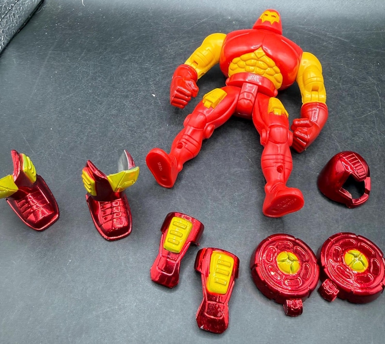 May include: A red and yellow Iron Man action figure with detachable parts. The figure is in a dynamic pose, with various components like the helmet, shoulder pads, and leg armor scattered around. The figure's design features a metallic finish.