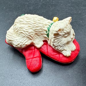 May include: A cream-colored cat figurine resting on a red stocking-shaped pillow. The cat has a green collar with a gold bell. The pillow has a quilted pattern. A festive Christmas decoration.