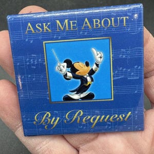 May include: A blue square button with a gold border and the text "Ask Me About By Request" in gold lettering. The button features a cartoon image of Mickey Mouse dressed in a tuxedo and conducting an orchestra.