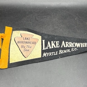 Könnte beinhalten: Schwarze Wimpel mit dem Text "Lake Arrowhead Myrtle Beach, S.C." und einem Logo mit der Aufschrift "Lake Arrowhead By The Sea". Die Wimpel hat eine gelbe Stoffschlaufe und Zierleiste.
