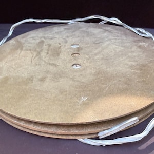 Op de afbeelding: Een stapel van drie cirkelvormige, lichtbruine houten schijven met een glad, plat oppervlak. Elke schijf heeft drie kleine, gelijkmatig verdeelde cirkelvormige gaten die verticaal in het midden zijn uitgelijnd. Een dunne, zilverkleurige draad loopt om de randen.