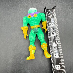 Op de afbeelding: Een groen en geel actiefiguur met een doorzichtige, koepelvormige kop en paarse accenten. De figuur is ongeveer 10 cm hoog, met gele laarzen en handschoenen. Een zwart en zilver meetlint ligt naast de figuur.