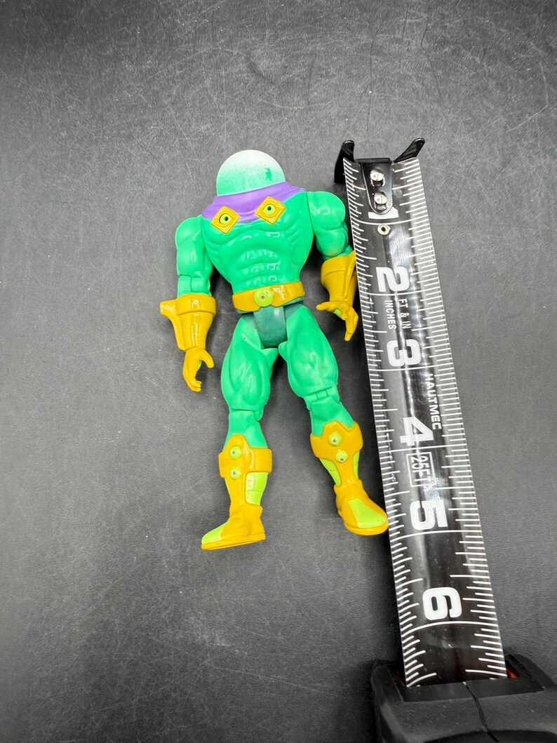 Op de afbeelding: Een groen en geel actiefiguur met een doorzichtige, koepelvormige kop en paarse accenten. De figuur is ongeveer 10 cm hoog, met gele laarzen en handschoenen. Een zwart en zilver meetlint ligt naast de figuur.