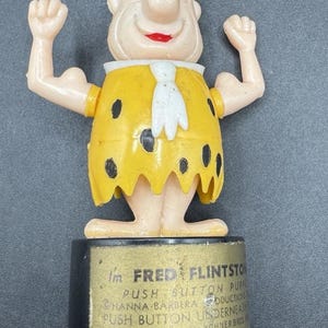 May include: A vintage Fred Flintstone push button puppet toy. The figure is wearing a yellow outfit with black spots and a white tie. The base is black with text that reads "I'm FRED FLINTSTONE PUSH BUTTON PUPPET".