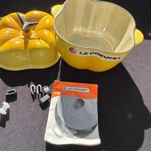 Puede incluir: Una fuente de cerámica amarilla Le Creuset con una tapa a juego en forma de pimiento. El plato tiene un interior blanco y asas laterales. También hay un pequeño folleto de instrucciones y varios accesorios pequeños en blanco y negro.