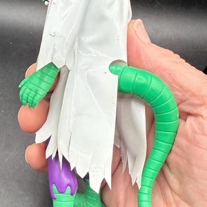 K&ouml;nnte beinhalten: Eine gr&uuml;ne und lila Actionfigur mit einem wei&szlig;en, zerfetzten Umhang. Die Figur hat einen langen gr&uuml;nen Schwanz und gr&uuml;ne H&auml;nde und F&uuml;&szlig;e. Die Figur wird in der Hand einer Person gehalten.