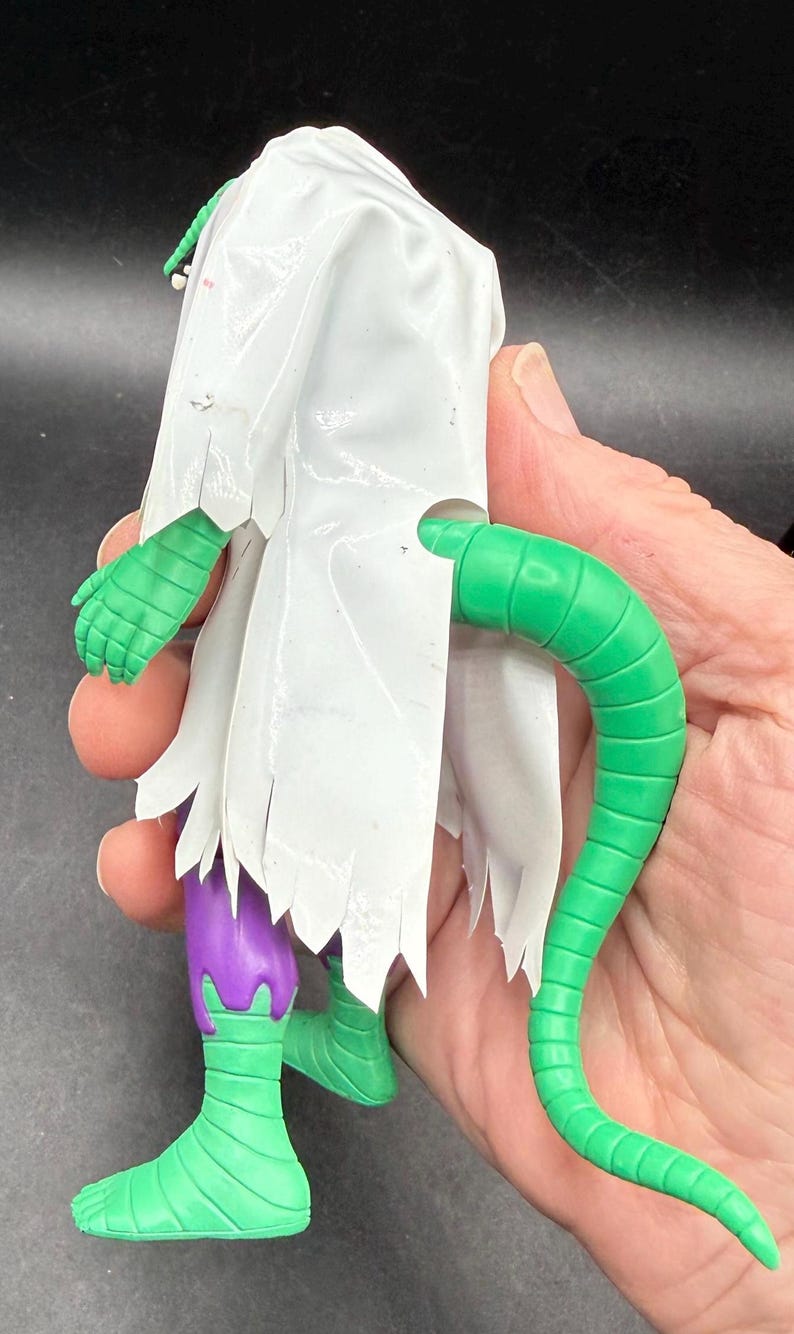 K&ouml;nnte beinhalten: Eine gr&uuml;ne und lila Actionfigur mit einem wei&szlig;en, zerfetzten Umhang. Die Figur hat einen langen gr&uuml;nen Schwanz und gr&uuml;ne H&auml;nde und F&uuml;&szlig;e. Die Figur wird in der Hand einer Person gehalten.