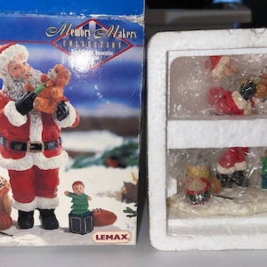Pode incluir: Um conjunto de duas figuras de Papai Noel em caixa, uma segurando um urso de pelúcia e a outra um tambor de brinquedo. A caixa é branca com detalhes vermelhos e azuis e o texto "Memory Makers Collection" impresso na frente.