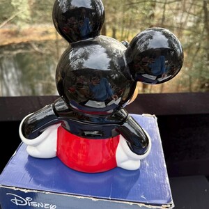May include: A ceramic Mickey Mouse figurine with a glossy black head and ears, a black jacket, red shorts, and white gloves. The figurine is sitting on a blue box with the Disney logo.