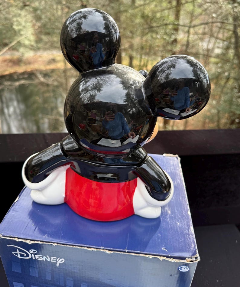 May include: A ceramic Mickey Mouse figurine with a glossy black head and ears, a black jacket, red shorts, and white gloves. The figurine is sitting on a blue box with the Disney logo.