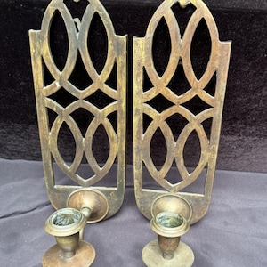May include: A pair of matching brass wall sconces featuring an elongated, pointed arch backplate with an intricate, openwork geometric lattice pattern. Each sconce includes a circular candle cup base extending outward from the lower section.