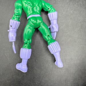 May include: A green action figure with a purple head, gloves, boots, and belt. The figure is posed with its back facing the viewer, showcasing the sculpted details of its muscles and costume. The figure's arms are bent, and it appears to be in a fighting stance.