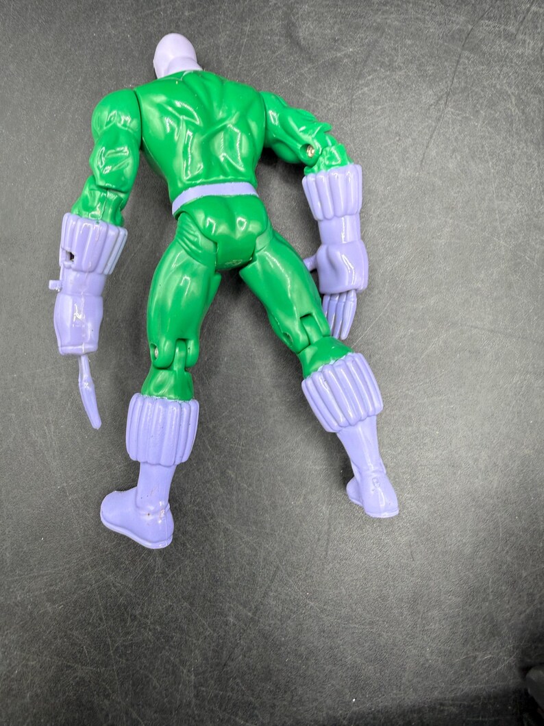 May include: A green action figure with a purple head, gloves, boots, and belt. The figure is posed with its back facing the viewer, showcasing the sculpted details of its muscles and costume. The figure's arms are bent, and it appears to be in a fighting stance.