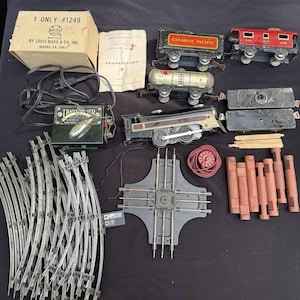 May include: A collection of vintage toy train set components, including a box labeled "1 ONLY #1249" and a transformer. The set features train cars with "CANADIAN PACIFIC" markings, tracks, and wooden supports. The items are arranged on a dark surface.