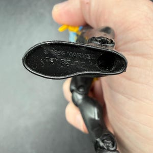 May include: A black action figure with yellow and blue accents. The underside of the foot is visible, with the text "&copy; 1995 MARVEL TOY BIZ INC." embossed on the sole. The figure is articulated.