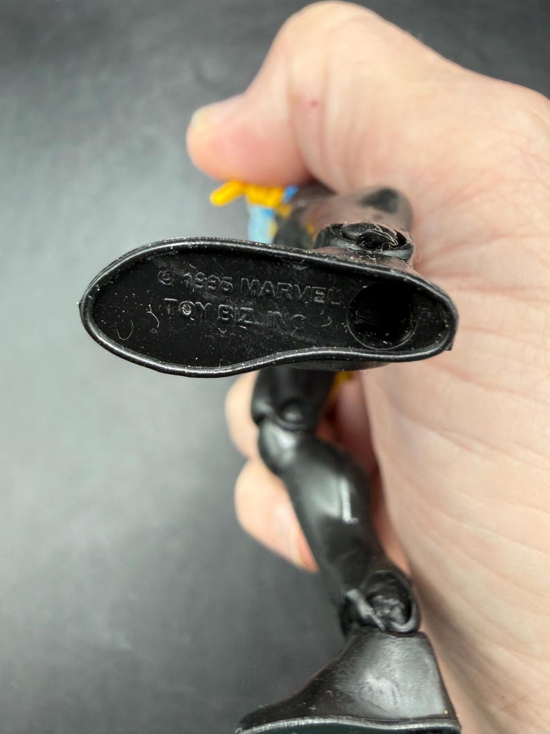 May include: A black action figure with yellow and blue accents. The underside of the foot is visible, with the text "&copy; 1995 MARVEL TOY BIZ INC." embossed on the sole. The figure is articulated.