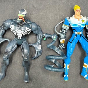 Op de afbeelding: Twee actiefiguren: een gespierde, donkergrijze Venom-figuur met witte accenten en een blauw-witte superheldenfiguur. Een grijs slangachtig accessoire staat ertussen. De figuren staan op een grijze ondergrond.