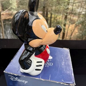 May include: A ceramic Mickey Mouse figurine, featuring a black head and ears, a tan face, and a red and white outfit. The figurine is posed on top of a blue box with the Disney logo.