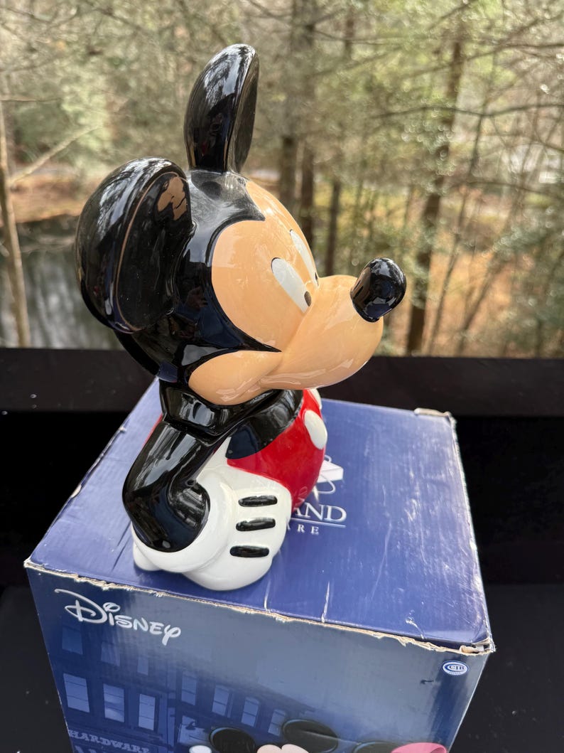 May include: A ceramic Mickey Mouse figurine, featuring a black head and ears, a tan face, and a red and white outfit. The figurine is posed on top of a blue box with the Disney logo.