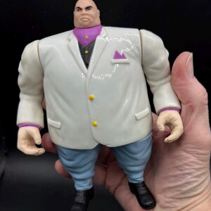 May include: A Kingpin action figure, featuring a white suit with yellow buttons, a purple ascot, and light blue pants. The figure has black shoes and a stern expression. The figure is held in a hand.