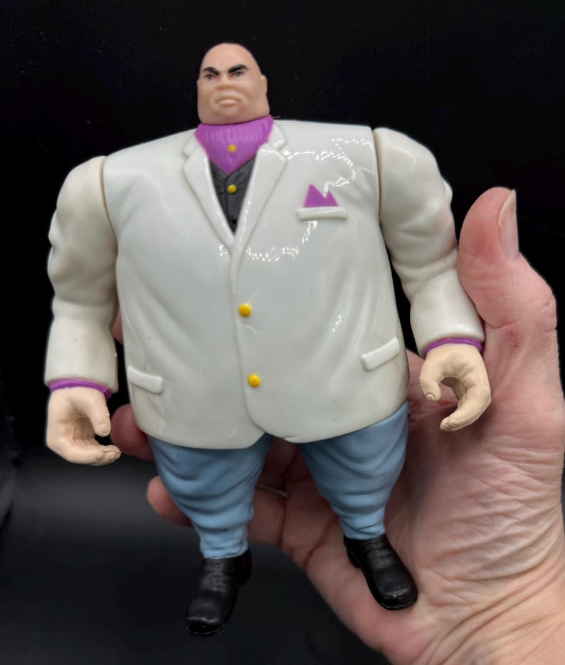 May include: A Kingpin action figure, featuring a white suit with yellow buttons, a purple ascot, and light blue pants. The figure has black shoes and a stern expression. The figure is held in a hand.