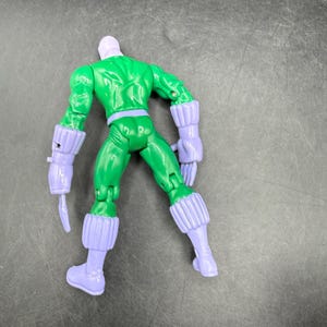May include: A green action figure with a purple head, gloves, and boots. The figure is posed with its back to the viewer, showcasing its muscular physique and detailed design. The figure has a belt and a weapon attached to its arm.