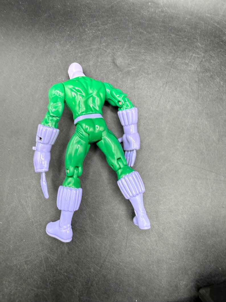 May include: A green action figure with a purple head, gloves, and boots. The figure is posed with its back to the viewer, showcasing its muscular physique and detailed design. The figure has a belt and a weapon attached to its arm.