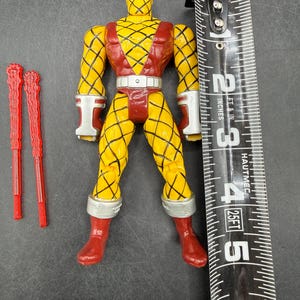 以下が含まれることがあります： 黄色と赤のアクションフィギュア。ダイヤモンド模様、赤いブーツ、白いアクセントが特徴です。フィギュアの高さは約13cmです。2つの赤いアクセサリーが含まれています。