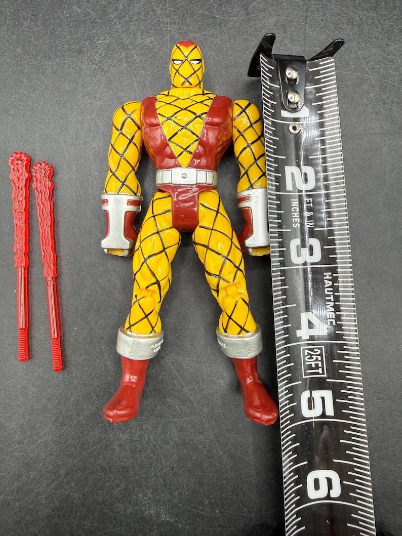 以下が含まれることがあります： 黄色と赤のアクションフィギュア。ダイヤモンド模様、赤いブーツ、白いアクセントが特徴です。フィギュアの高さは約13cmです。2つの赤いアクセサリーが含まれています。