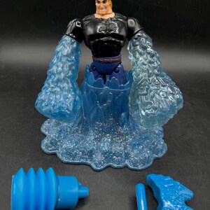 May include: A superhero action figure with a black top, blue pants, and blonde hair. The figure has translucent blue water-like arms and a base. Two blue accessories are also included.