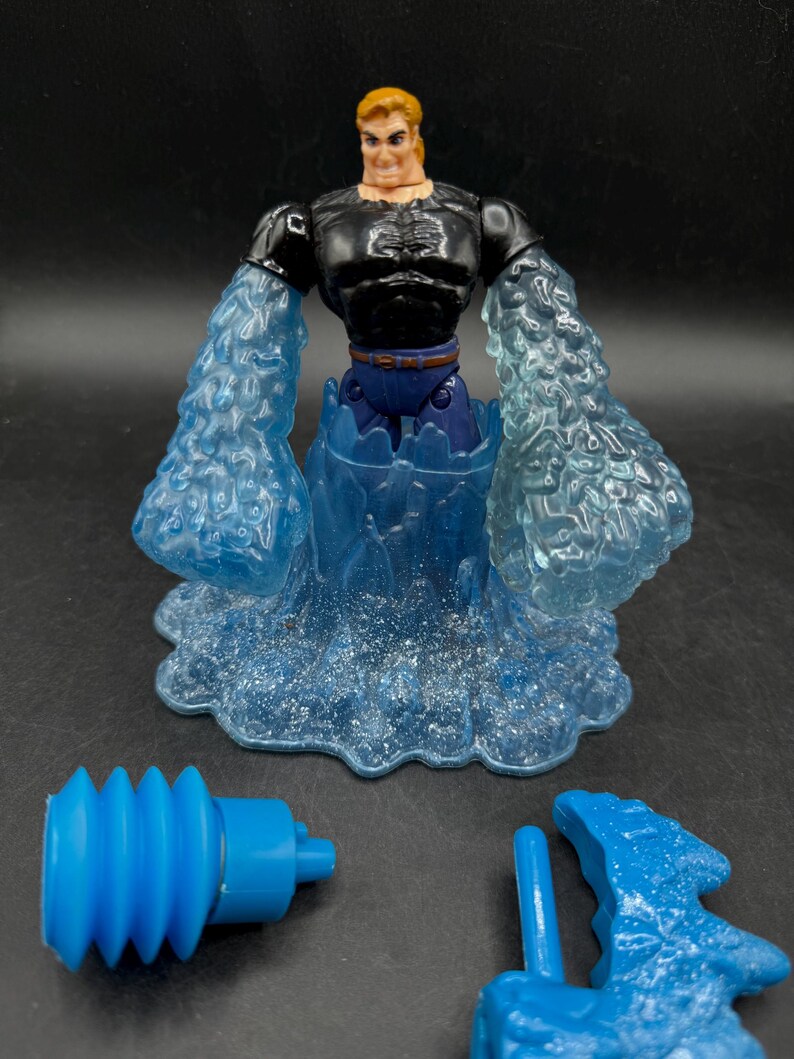 May include: A superhero action figure with a black top, blue pants, and blonde hair. The figure has translucent blue water-like arms and a base. Two blue accessories are also included.